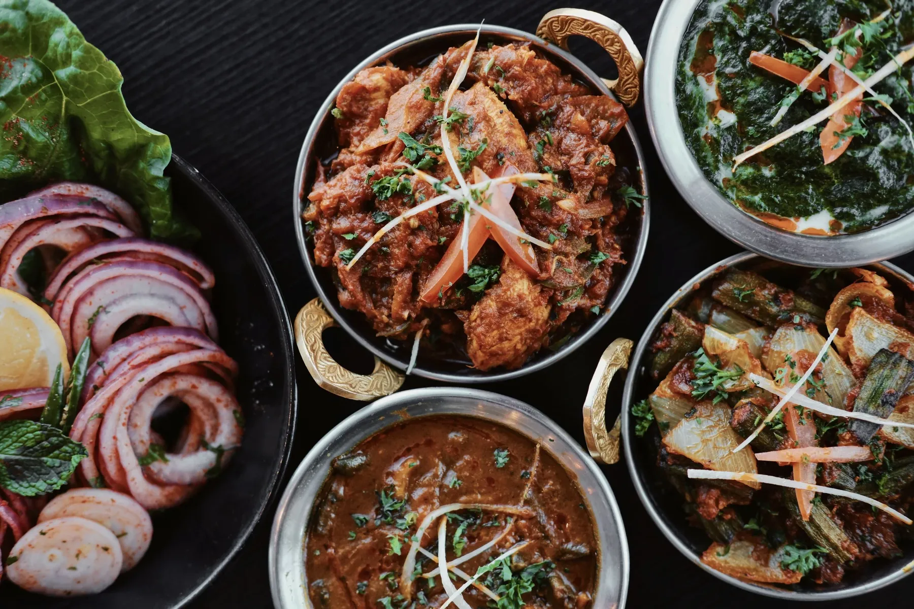 Curries in copper karahis, masala onion salad and palak paneer on a dark table at Taste of India