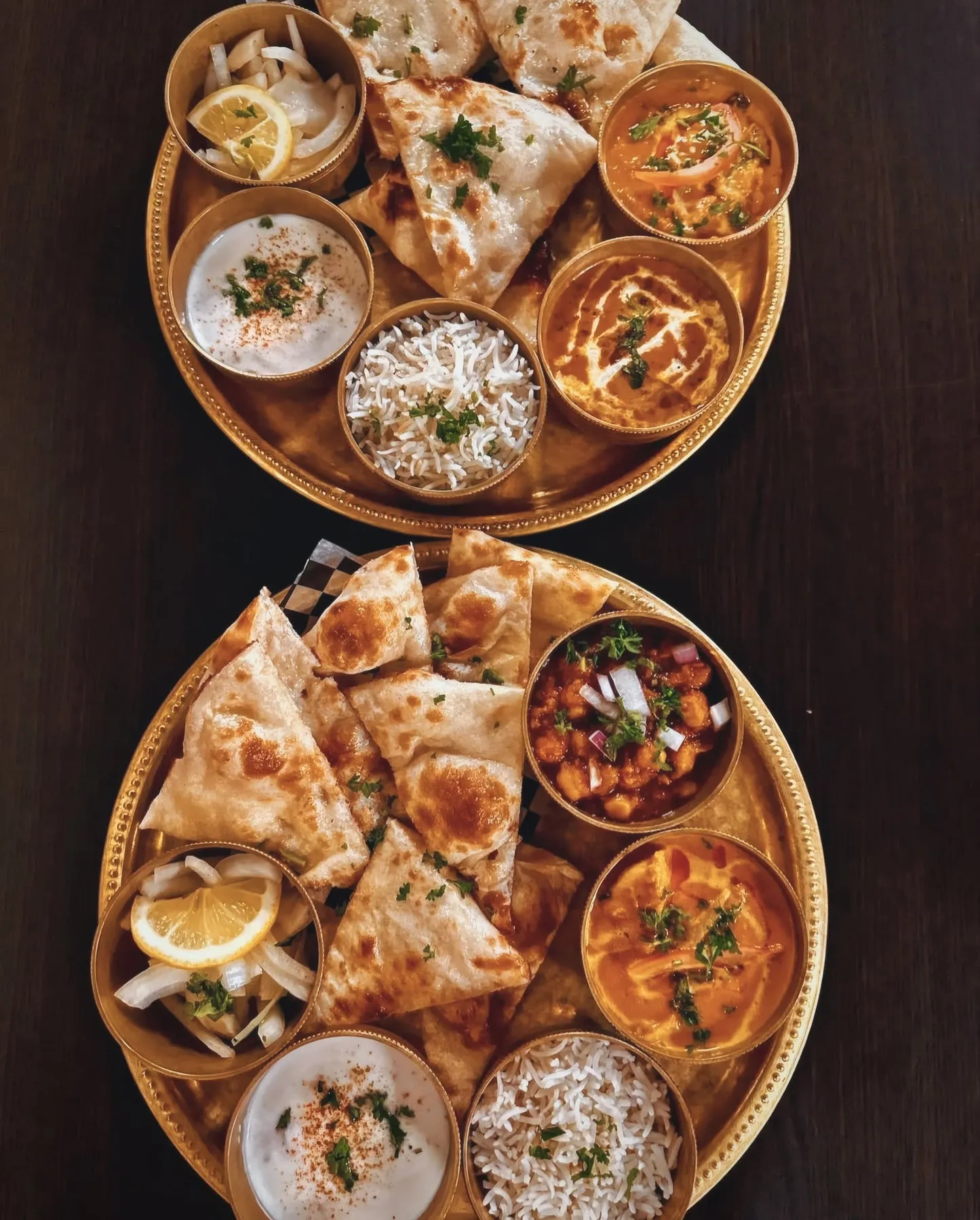 Two gold thali platters with naan, samosas, curries, rice and raita